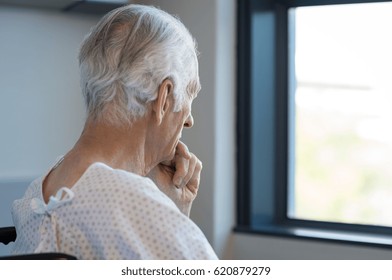 Rear View Of A Senior Man Sitting On Wheelchair Looking Outside The Window. Old Man In Hospital Room Sitting Near Window And Thinking. Elderly Patient Feeling Sad And Alone At Hospital.