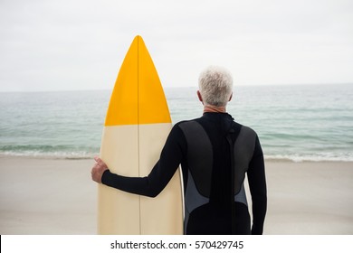 Rear view of senior man in wetsuit holding a surfboard on the beach - Powered by Shutterstock