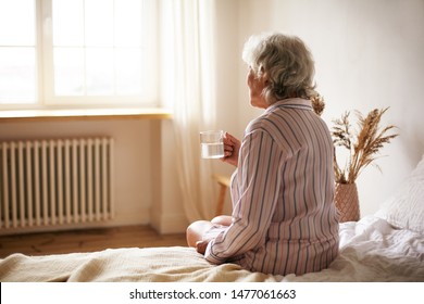 Rear View Of Senior Sixty Year Old Woman With Gray Hair Holding Mug Washing Down Sleeping Pill, Suffering From Insomnia. Elderly Retired Female Taking Medicine With Water, Sitting In Bedroom