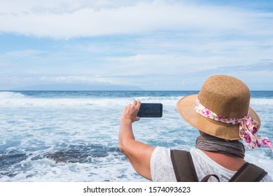 Rear View Of A Senior Woman At The Sea In Winter Time Taking A Photo Of The Horizon Over Water. Casual Clothing With Backpack And Hat. Cloudy Sky