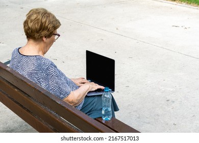 Rear View At Senior Woman Sitting On Park Bench Using On Laptop.