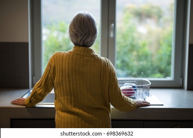 Rear view of senior woman standing near the kitchen sink and looking through window