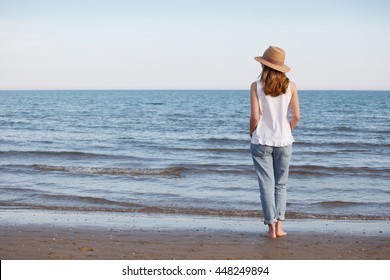Rear view shot of a barefoot mature woman standing along a sandy beach while wearing straw hat and casual clothes.