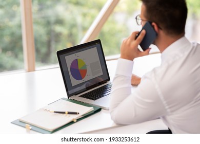 Rear view shot of financial advisor businessman talking on his mobile phone and using laptop while working at office desk. - Powered by Shutterstock