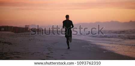Rear view shot of fit man running on the sea shore in morning. Healthy male athlete running along the beach.