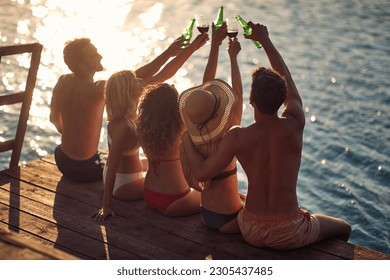 Rear view shot of group of young people sitting on wooden jetty by water, cheering with drinks. Couples on vacation. Holiday, togetherness, lifestyle concept.