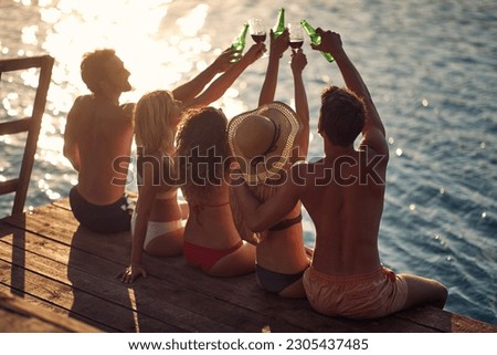Rear view shot of group of young people sitting on wooden jetty by water, cheering with drinks. Couples on vacation. Holiday, togetherness, lifestyle concept.