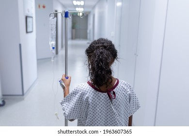 Rear View Of Sick Mixed Race Girl Walking In Hospital Corridor With Iv Drip Bag On Stand. Medicine, Health And Healthcare Services.