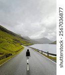 Rear view of a single female walking on an empty road surrounded by lush green mountains, fjords and the sea. She is dressed casually in hiking gear. Eysturoy, Faroe Islands, Denmark.