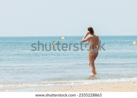Rear view of a single lady walking into the sea wearing a bikini with a thong; back view of a single female paddling in a calm, azure sea wearing a light blue bikini with a thong

