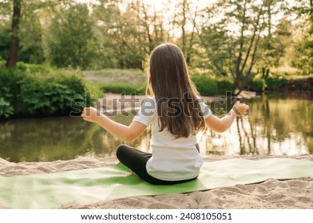 Rear view of small girl with brown flowing hair calmly sitting on green mat on sandy plage and doing yoga pose. Little athlete going for sports and meditating in front of lake at sunset.