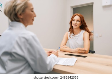 Rear view of smiling caucasian woman doctor listening young woman patient complaints, making notes in card clipboard or healthcare insurance form, at initial medical consultation appointment - Powered by Shutterstock