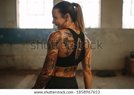 Rear view of sportswoman smiling after workout session. Tattooed woman in sportswear taking break after shadow boxing exercise.