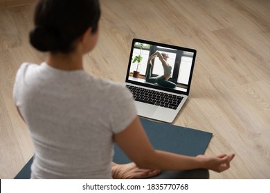 Rear view sporty Indian young woman practicing yoga at home, sitting in lotus pose on mat on floor, watching webinar online, using laptop, doing exercise, stretching, enjoying fitness activity