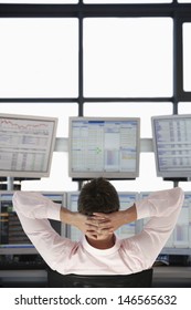 Rear view of stock trader with hands behind head watching multiple monitors