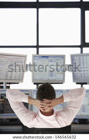 Rear view of stock trader with hands behind head watching multiple monitors