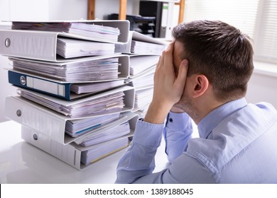 Rear View Of Stressed Businessman With Hand On Head In Front Of Stacked Folders