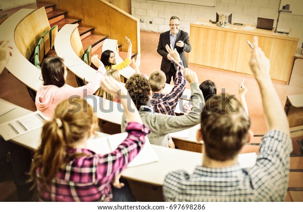 Rear view of students raising hands with teacher in\
college lecture hall