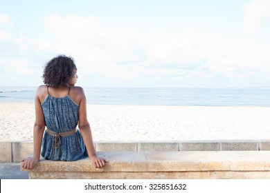 Rear view of a stylish african american young woman sitting by white sand beach contemplating the sea on a sunny summer holiday destination, outdoors. Travel lifestyle and healthy living, exterior.