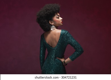 Rear View Of A Stylish Young African American Woman In An Elegant Green Evening Dress Against Maroon Background.