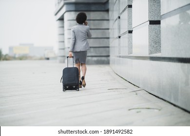 Rear view of a successful black business woman walking with her luggage and using smart phone outside of the airport.