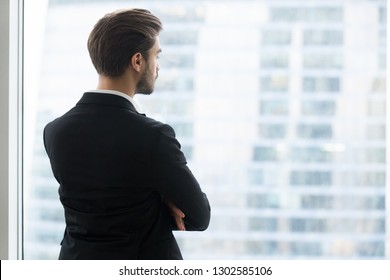 Rear View At Successful Businessman Looking Through Window At Modern City Skyscrapers Thinking About Future Opportunity, Planning New Goals, Making Decision, Business Vision And Peace Of Mind Concept