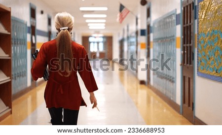 Rear view of teacher walking down an empty school hallway holding books with US American flag in the distance with copy space.