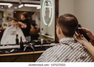 Rear View Of A Teenager Boy Having His Hair Cut With A Trimmer At The Barber Shop. Hair Care Service Concept. Blurred Reflection In A Mirror. Focus On A Head.