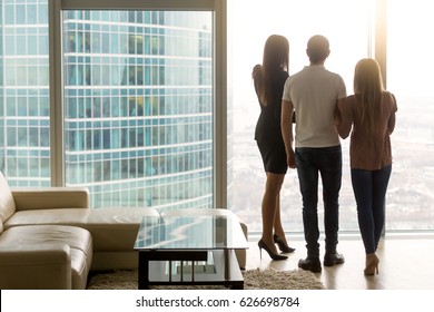 Rear View Of Three Business People Standing By The Full Length Floor-to-ceiling Window In Office, Looking At Big City Scenery. Real Estate Agent Showing Apartment To Couple, Enjoying Birds-eye View 