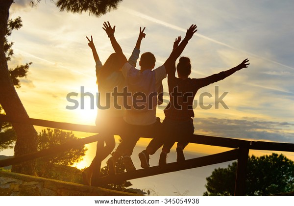 Rear view of three couple best friends travelers\
put hands up at sunset. Young  relaxing hipster wanderers enjoying\
exclusive alternative destination. Holiday life moment at warm\
orange light