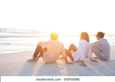 Rear view of three friends men sitting together on a white sand beach with the sun setting behind them with warm orange light, during their vacation in an idyllic nature scene destination. - Powered by Shutterstock