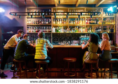 Rear view of three guys drinking beer, looking at women, two girlfriends sitting at the bar counter. Friends spending time at night club, restaurant. Horizontal shot