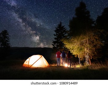 Rear View Three Person Tourists Looking At The Burning Fire Near The Glowing Orange Tent On One Side And The Trees On The Other Under The Unusual Starry Sky And Milky Way On Background Of Mountains.