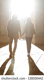 Rear View Of A Tourist Couple Silhouette Holding Hands While Visiting A Destination City, Walking Under An Old Arch With The Morning Sun Rays Filtering Through.