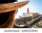 Rear view of a tourist woman wearing a hat, enjoying the breathtaking view of Farol do Cabo de São Vicente lighthouse perched on the rugged cliffs of cape São Vicente, a must-visit tourist destination