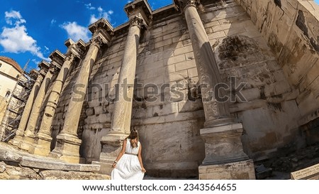 Rear view of tourist woman wearing white dress and golden laurel crown walking through ruined Library of Hadrian in Athens, Attica, Greece, Europe. Corinthian columns of propylon of Pentelic marble