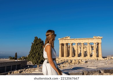 Rear View Of Tourist Woman In White Dress Looking At Parthenon Of Acropolis Of Athens, Attica, Greece, Europe. Ruins Of Ancient Temple, The Birthplace Of Democracy. Girl Wearing Golden Laurel Crown