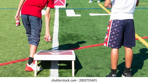Rear View Of Two Boys Playing Cornhole Bean Bag Toss Game On A Turf Field.