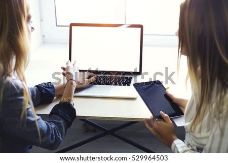 Rear view of two female IT developer testing usability of software application for touch pad using wireless connection to internet and computer laptop with mock up screen sitting in coworking space