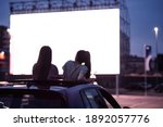 Rear view of two female friends sitting in the car while watching a movie in an open air cinema with a big white screen. Entertainment concept. Focus on people. Horizontal shot