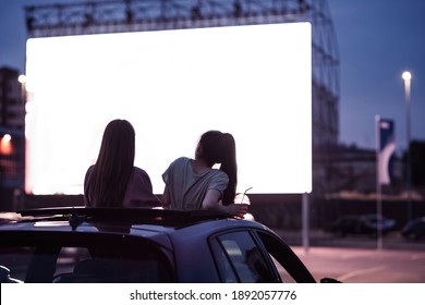 Rear View Of Two Female Friends Sitting In The Car While Watching A Movie In An Open Air Cinema With A Big White Screen. Entertainment Concept. Focus On People. Horizontal Shot