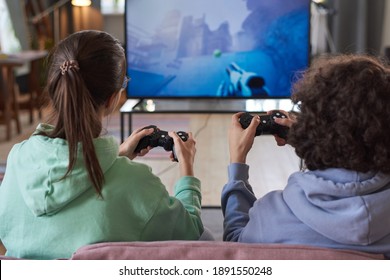 Rear View Of Two Friends Playing Video Game In Team Sitting On Sofa In Front Of Big TV