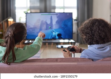 Rear View Of Two Friends Sitting On Sofa And Playing Video Game Together On Big TV