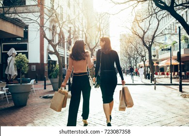 Rear View Of Two Friends Walking On The City Street With Shopping Bags. Female Shoppers Carrying Shopping Bags While Walking Along The Road.