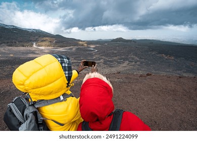 Rear view of two hikers in colorful jackets taking photo of dramatic mountain scenery with smartphone. Depicts adventure travel, outdoor photography, and technology use in nature. - Powered by Shutterstock