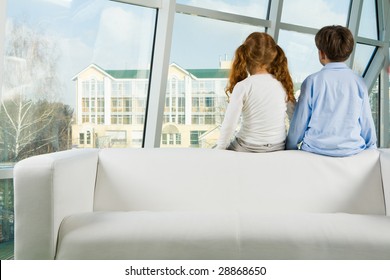 Rear View Of Two Kids Sitting On White Sofa And Looking Through Window