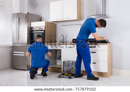Rear View Of Two Men Checking Induction Stove And Sink Pipe In Kitchen