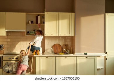 Rear view of two naughty kids, brother and sister trying to find something sweet in the kitchen cabinet. Little girl helping her brother by holding his hand. Childhood, children safety concept