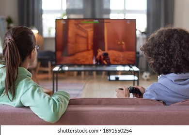 Rear View Of Two Teenagers Sitting On Sofa In Front Of Big Screen Of TV And Playing Video Game At Home