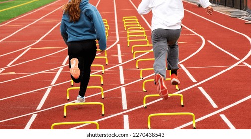 Rear view of two track athletes running over small yellow hurdles in lane on a track in the cold at practice. - Shutterstock ID 2270438543